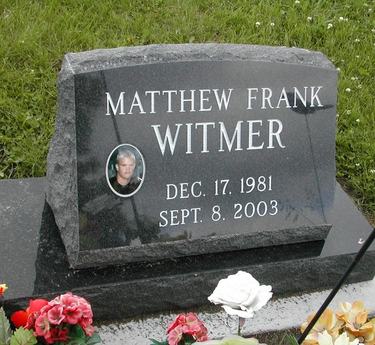 Black granite slant marker on a base with crisp engraved lettering, an oval photo, and flowers in a cemetery lawn.
